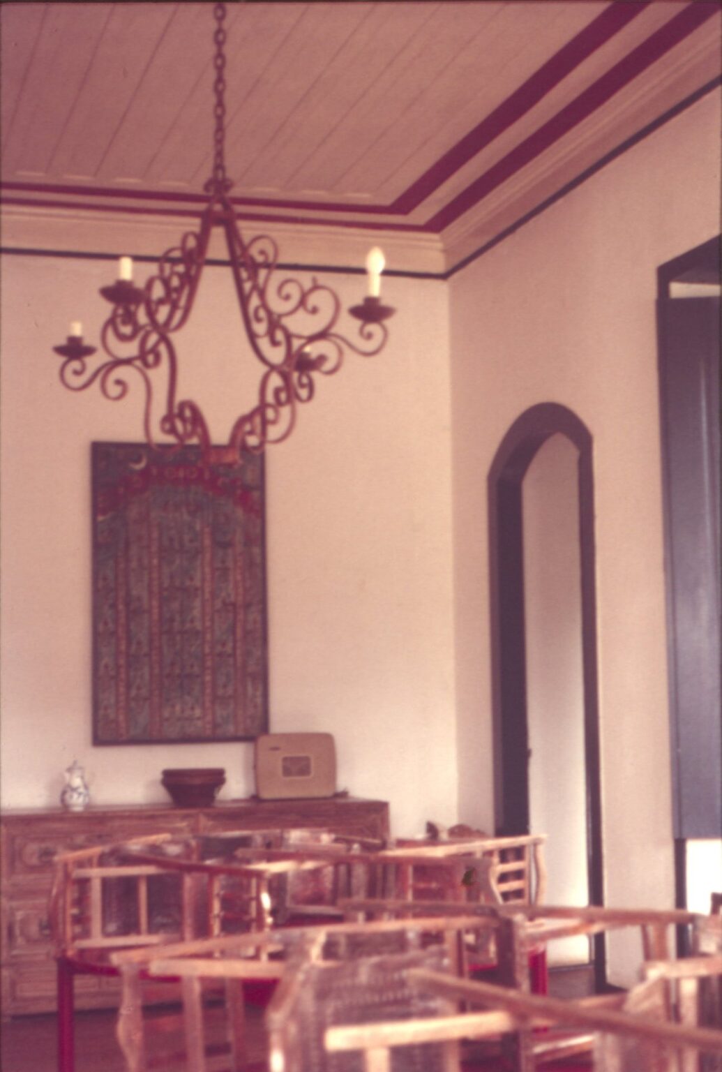 Vista interna de salão com lustre de ferro, tapeçaria na parede e mobiliário de madeira — P0024 — Fazenda Fernando Millan — F016-P0024-1981-S03-D00019