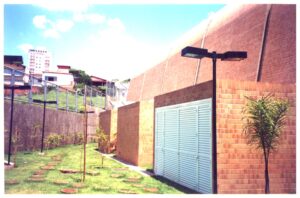Vista lateral de edifício com cobertura abobadada revestida em telhas cerâmicas, jardim gramado com pedras de passagem e postes de iluminação, ao fundo prédios e casas da vizinhança — P0131