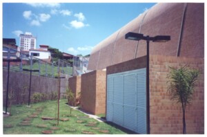 Vista lateral de edifício com cobertura abobadada revestida em telhas cerâmicas, jardim gramado com pedras de passagem e postes de iluminação, ao fundo prédios e casas da vizinhança — P0131 — SESC - Piracicaba