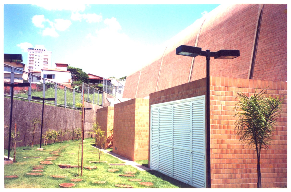 Vista lateral de edifício com cobertura abobadada revestida em telhas cerâmicas, jardim gramado com pedras de passagem e postes de iluminação, ao fundo prédios e casas da vizinhança — P0131 — SESC - Piracicaba — F009-P0131-0000-S03-D00052