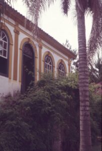 Vista parcial da fachada de edificação antiga, com portas e janelas em arco emolduradas, telhado colonial e vegetação (palmeira e arbustos) em primeiro plano — P0024 — Fazenda Fernando Millan