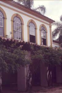 Vista parcial da fachada superior de edifício de arquitetura colonial, com janelas em arco emolduradas e varanda inferior coberta por vegetação florida — P0024 — Fazenda Fernando Millan